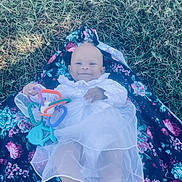 Haislyn is registered to the contest to win money with this photo: baby, child, smiling, white_dress, toy, colorful, grass, floral_blanket, outdoor, infant, cute, playing, lying_down, person, happy, nature, soft_light, small_toy, bare_foot, portrait