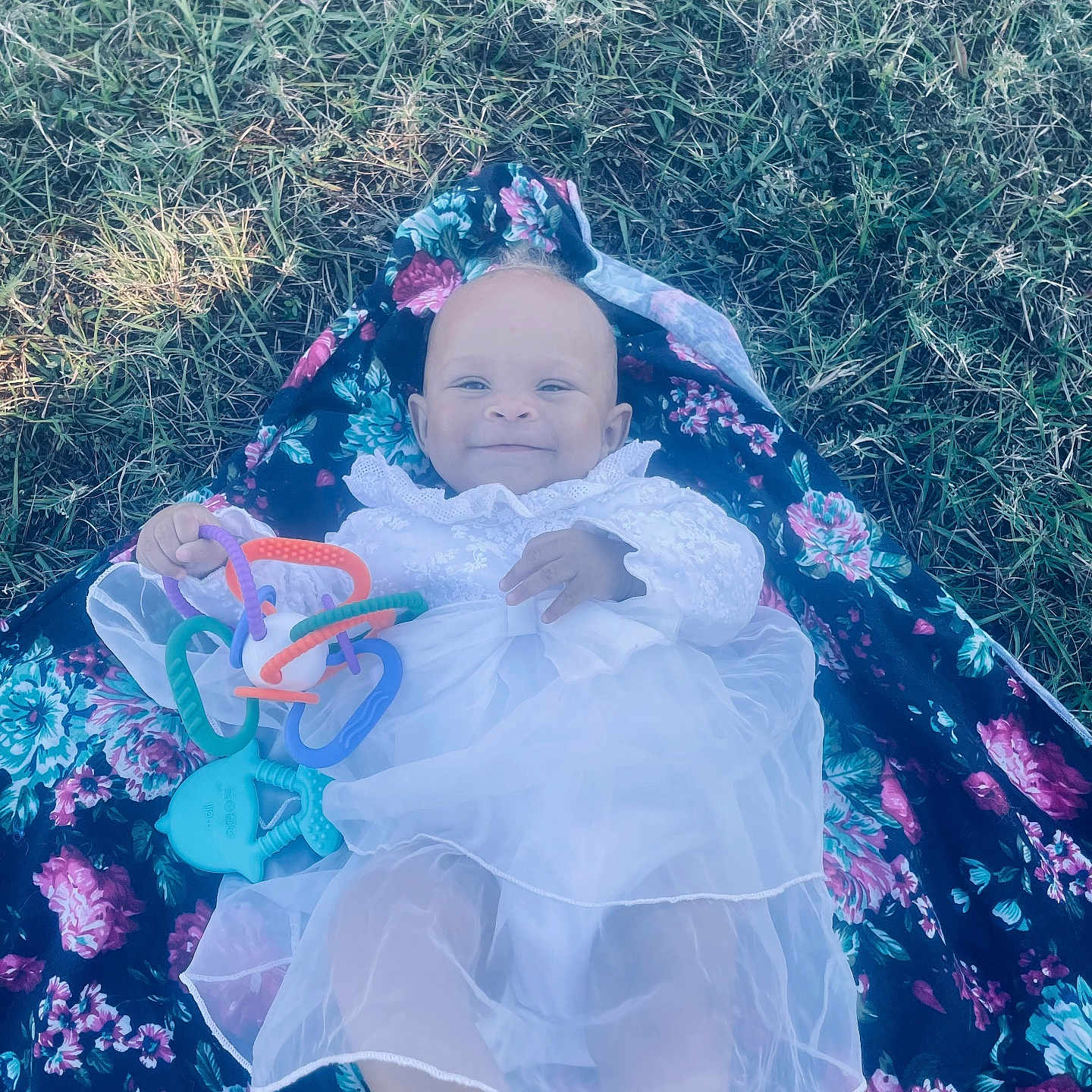 Haislyn is registered to the contest to win money with this photo: baby, bare_foot, child, colorful, cute, floral_blanket, grass, happy, infant, lying_down, nature, outdoor, person, playing, portrait, small_toy, smiling, soft_light, toy, white_dress