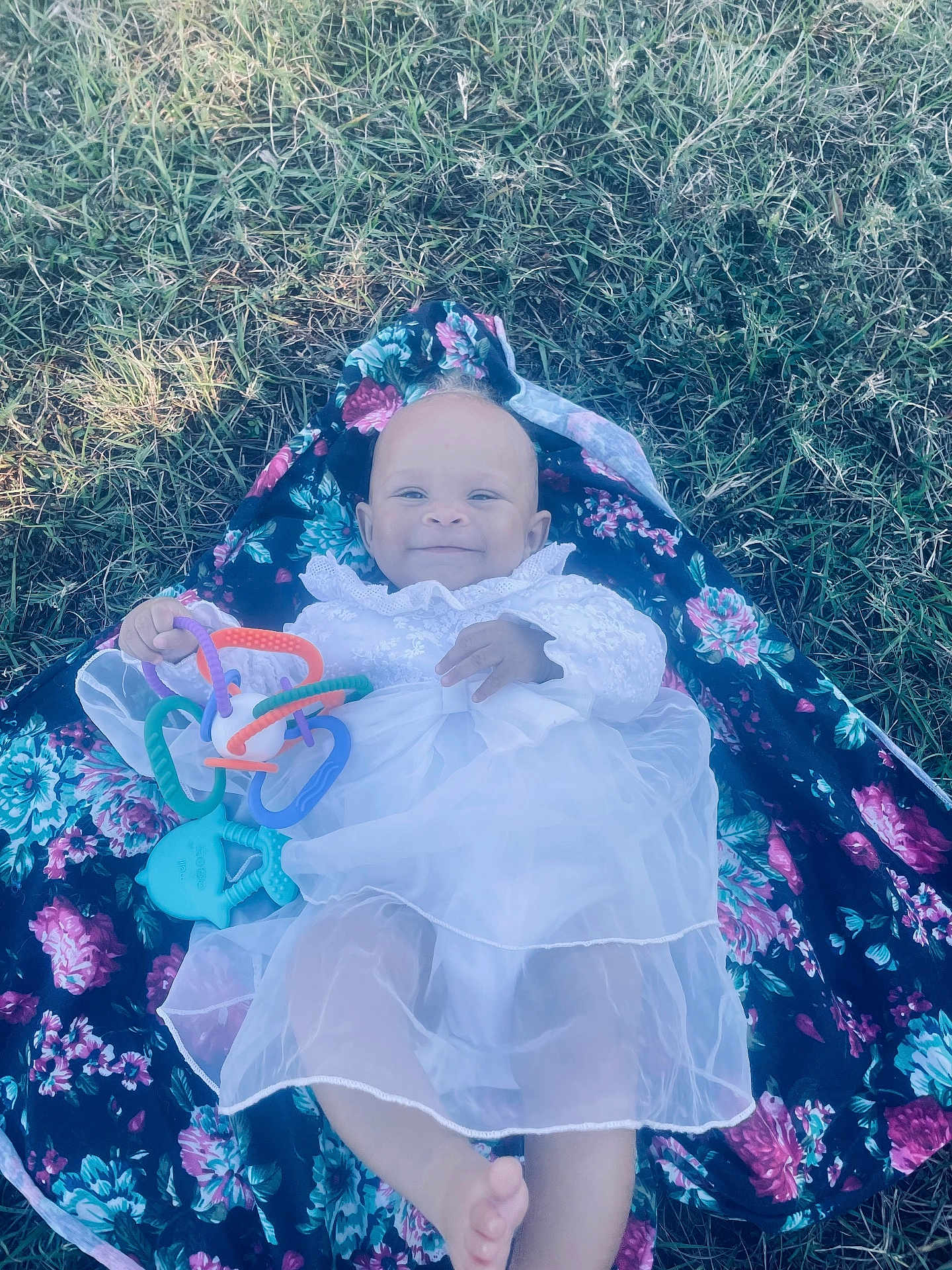 Haislyn is registered to the contest to win money with this photo: baby, child, smiling, white_dress, toy, colorful, grass, floral_blanket, outdoor, infant, cute, playing, lying_down, person, happy, nature, soft_light, small_toy, bare_foot, portrait