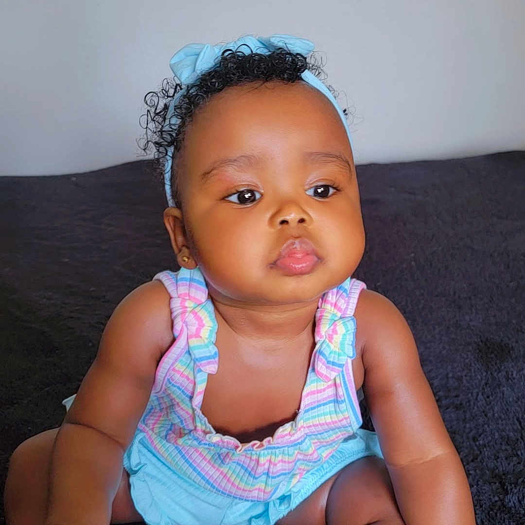 Ocean is registered to the contest to win money with this photo: baby, blue_headband, child, chubby_cheeks, curly_hair, cute, dark_background, expression, face, frills, headband, holding_foot, indoor, infant, pastel_colors, portrait, romper, sitting, skin, white_socks