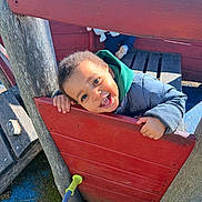 Ismaël participe au concours pour gagner de l'argent avec cette photo : child, toddler, smiling, playground, play_structure, wood, red_panel, green_hood, jacket, happy, face, portrait, outdoor, sunlight, hands, teeth, bench, shoe, playground_surface, toy_handle