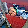 child, toddler, smiling, playground, play_structure, wood, red_panel, green_hood, jacket, happy, face, portrait, outdoor, sunlight, hands, teeth, bench, shoe, playground_surface, toy_handle