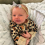 Halston is registered to the contest to win money with this photo: baby, infant, onesie, leopard_print, headband, bow, blanket, smile, face, hands, cozy, comfort, couch, cute, sleepwear, newborn, person, portrait, indoor, soft