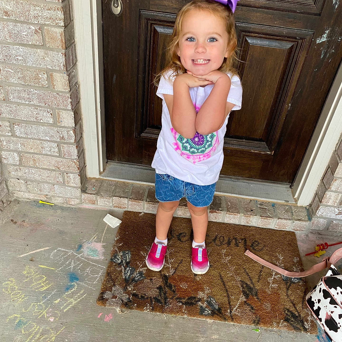 Brixlyn is registered to the contest to win money with this photo: brick_wall, casual_clothing, chalk_drawings, child, concrete_floor, daylight, denim_shorts, front_porch, girl, happy, outdoor, pink_shoes, playful, posing, purple_bow, smiling, welcome_mat, white_tshirt, wooden_door, young_child