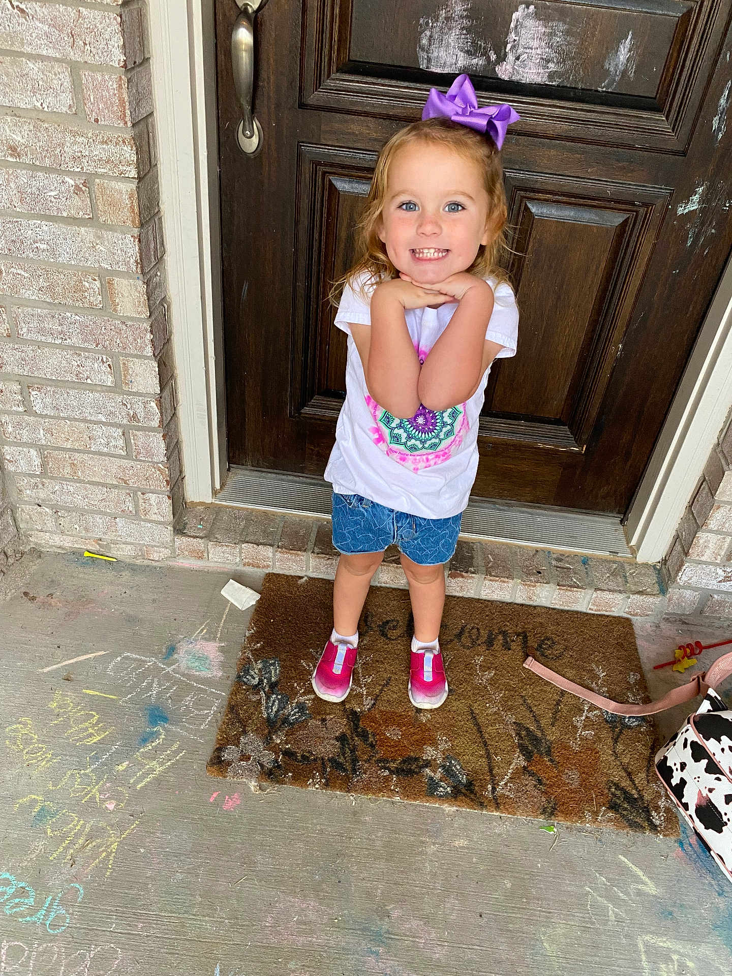 Brixlyn is registered to the contest to win money with this photo: child, girl, purple_bow, white_tshirt, denim_shorts, pink_shoes, welcome_mat, chalk_drawings, concrete_floor, wooden_door, brick_wall, smiling, posing, outdoor, front_porch, happy, casual_clothing, young_child, daylight, playful