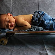 Ozzy is registered to the contest to win money with this photo: baby, newborn, sleeping, skateboard, jeans, clothing, soft_pillow, gray_background, infant, peaceful, portrait, cute, child, resting, floor, studio, casual, small, horizontal, toddler