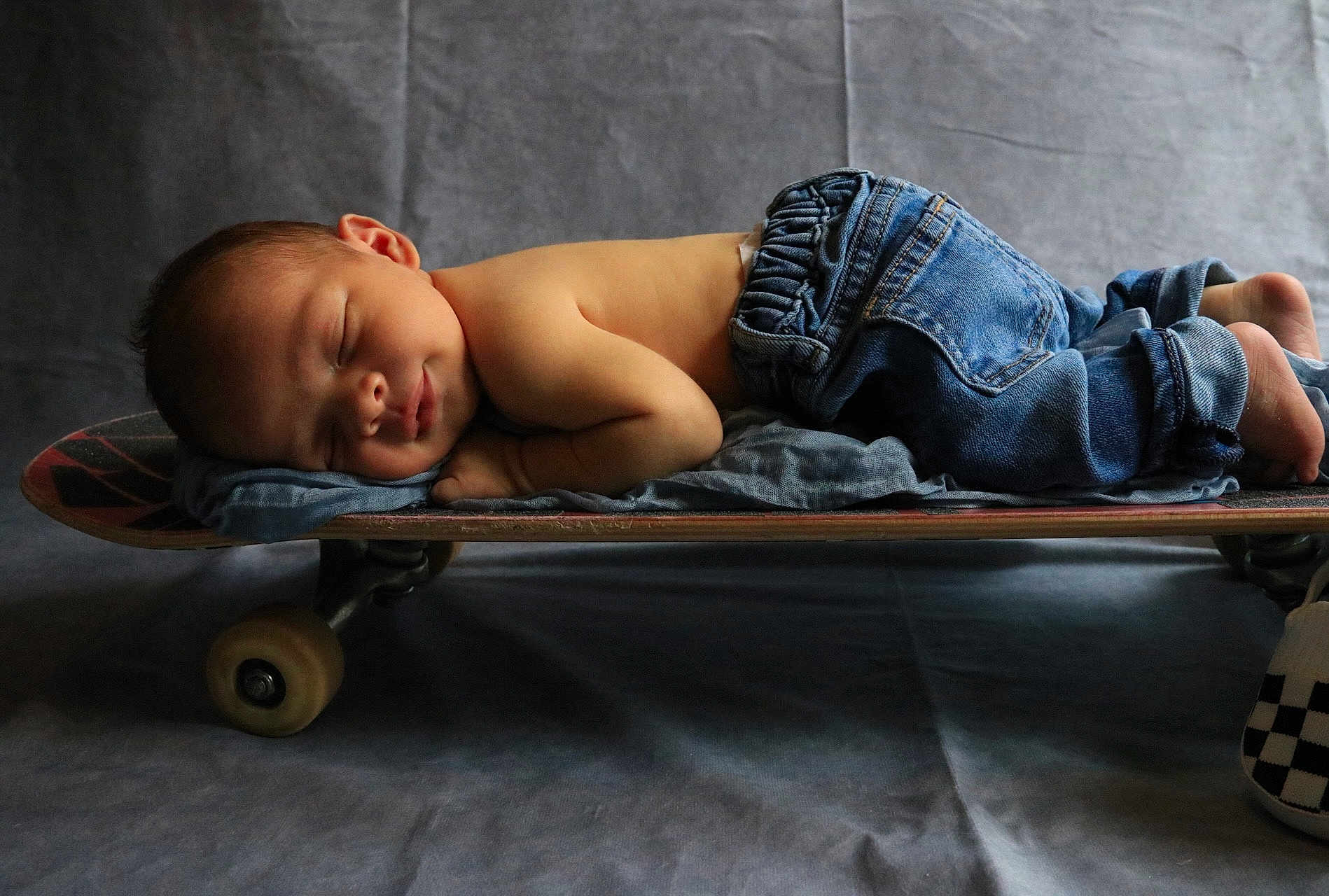 Ozzy is registered to the contest to win money with this photo: baby, newborn, sleeping, skateboard, jeans, clothing, soft_pillow, gray_background, infant, peaceful, portrait, cute, child, resting, floor, studio, casual, small, horizontal, toddler
