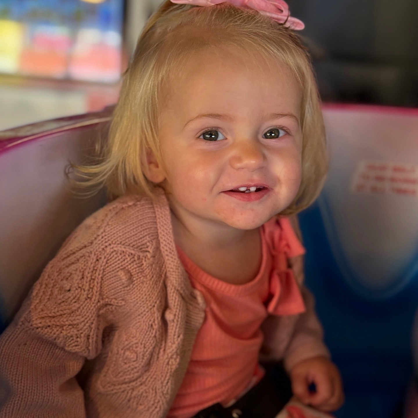 Amelia is registered to the contest to win money with this photo: blonde_hair, bright, child, clothing, colorful_seat, cute, face, fashion, happy, indoor, knitted_cardigan, person, pink_bow, playful, portrait, seatbelt, sitting, smile, toddler, young_child
