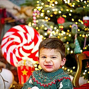 Ilyas a rejoint le concours — aidez-le/la à gagner de superbes lots ! child, toddler, smile, sweater, holiday, christmas_tree, lights, decorations, candy, popcorn, chair, indoors, festive, happy, winter, face, person, toy, background, celebration