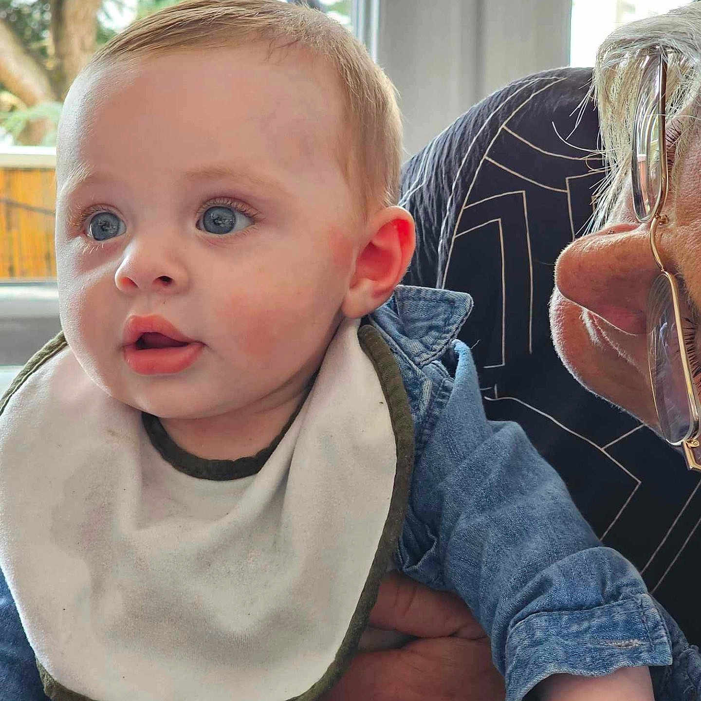 Gabriel participe au concours pour gagner de l'argent avec cette photo : adult, baby, bib, blue_eyes, child, close_up, curious, denim_jacket, expression, face, glasses, hand, holding, indoor, natural_light, person, short_hair, skin, table, window