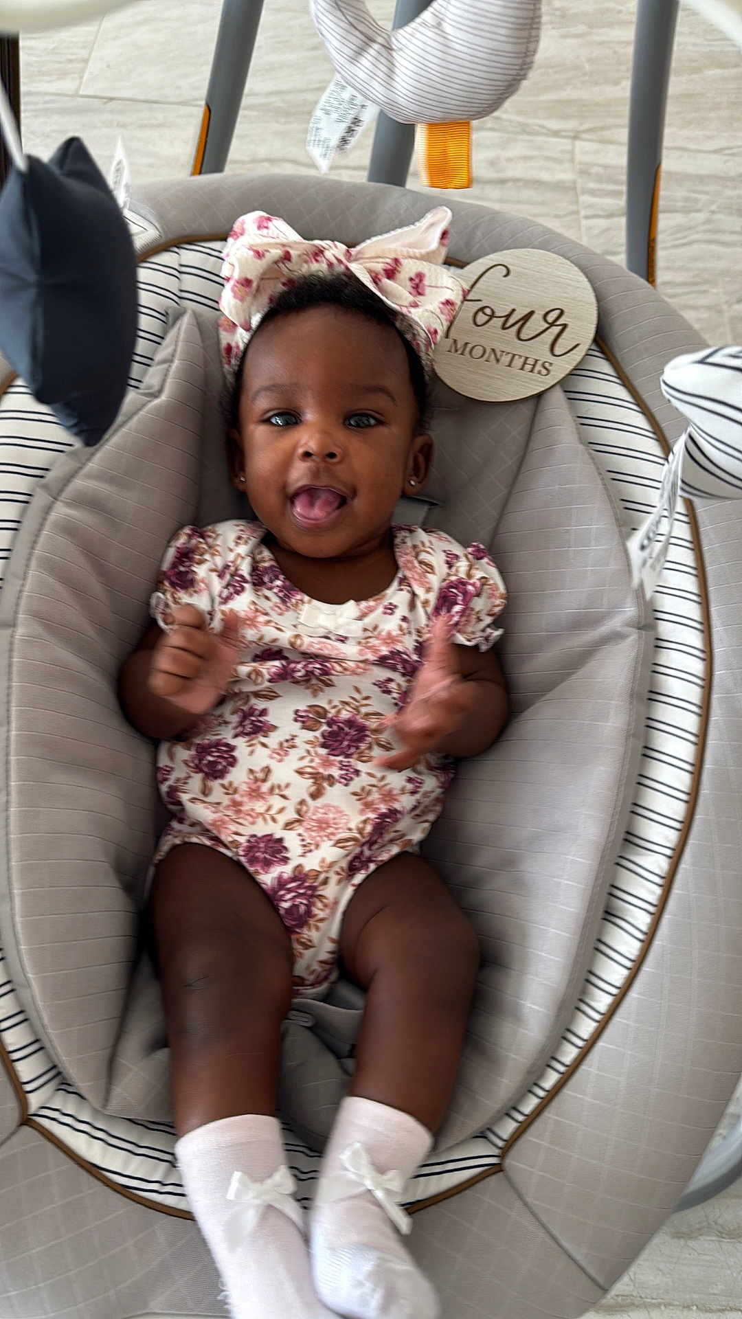 Khalani is registered to the contest to win money with this photo: baby, infant, child, floral_clothing, bow, baby_rocker, clapping, happy, smiling, socks, toy, indoor, person, cute, young_child, headband, seat, playful, comfortable, expression