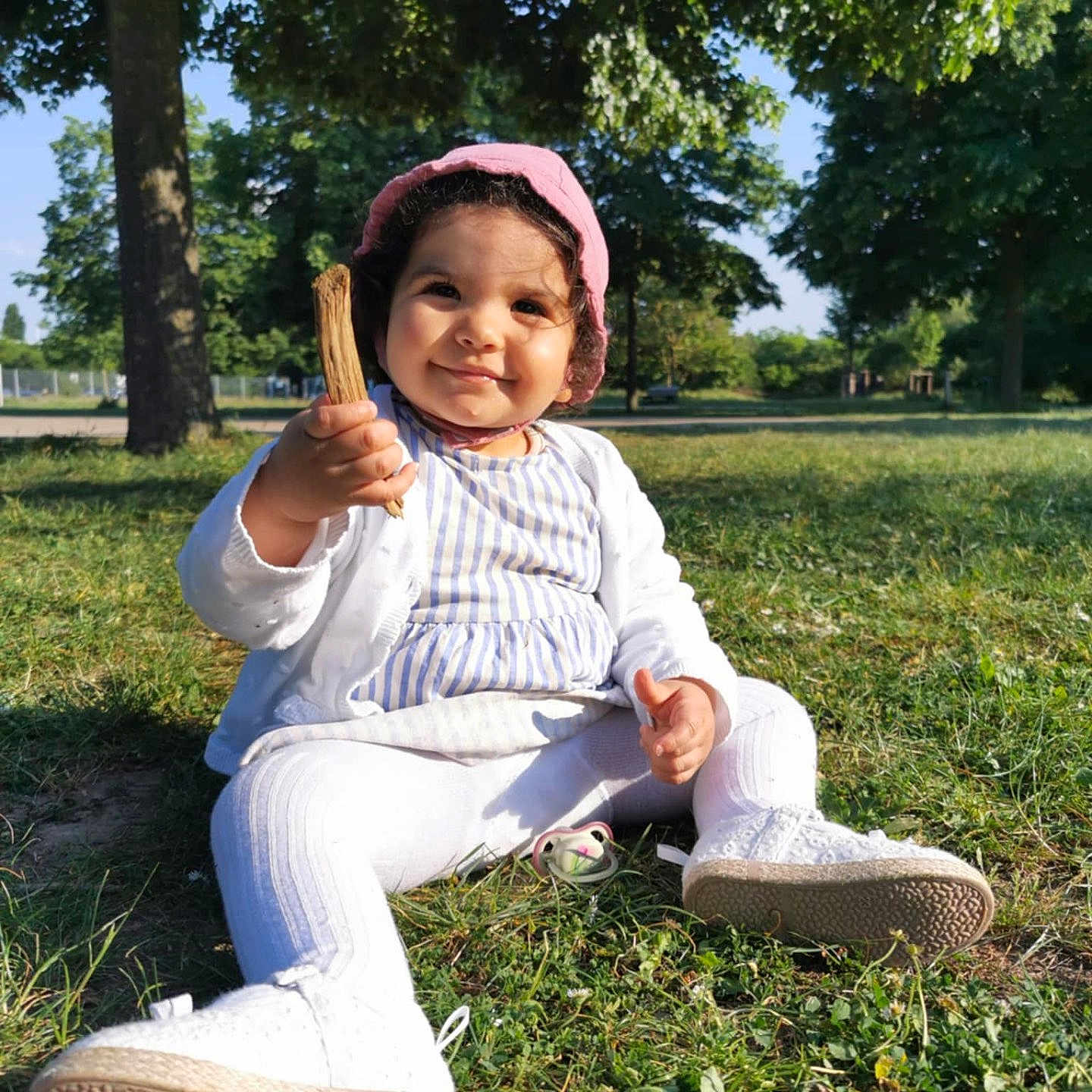 Elena participe au concours pour gagner de l'argent avec cette photo : toddler, child, grass, park, trees, sunlight, outdoor, pink_bonnet, white_cardigan, striped_dress, white_leggings, white_shoes, stick, pacifier, smiling, sitting, nature, daylight, cute, face