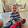 baby, blurred_background, chair, child, dining_room, eyes, hair, hands, happy, indoor, playtime, plush_rug, portrait, red_toy, ride_on_toy, smiling, striped_sweater, tile_floor, toddler, wheel