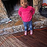 bow, casual_clothing, child, confident, cozy, cute, extension_cord, fireplace, girl, hands_behind_back, home, indoor, jeans, pink_sweatshirt, portrait, pout, sneakers, standing, stone_wall, wood_floor
