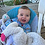 baby, smiling, blanket, baby_seat, outdoor, wooden_deck, flower, greenery, happy, child, cozy, blue_clothing, face, person, cute, infant, nature, daylight, comfort, portrait