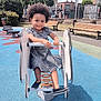Adr a rejoint le concours — aidez-le/la à gagner de superbes lots ! child, playground, spring_rocker, smiling, outdoor, curly_hair, dress, black_shoes, bench, rubber_floor, park, daylight, happy, person, recreation, fun, urban, greenery, fence, buildings