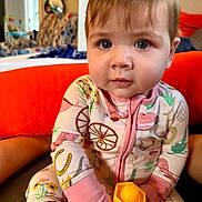 Emmy joined the competition — help win amazing prizes! baby, child, onesie, toy, yellow_toy, indoor, orange, face, hands, sitting, curious, patterned_clothing, furniture, couch, living_room, colorful, expression, young_child, portrait, closeup