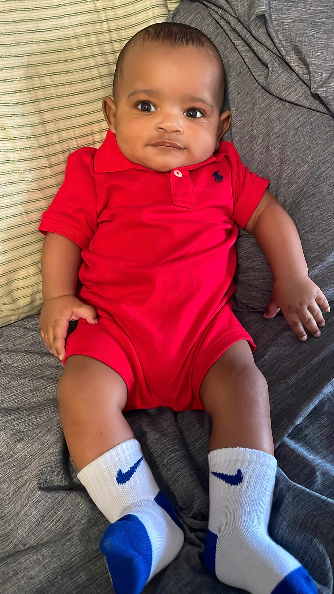 Jahlil joined the competition — help win amazing prizes! baby, infant, red_onesie, white_socks, blue_socks, nike_logo, couch, striped_pillow, indoor, child, portrait, smiling, cute, relaxed, sitting, clothing, feet, hand, face, young