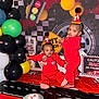 balloon, birthday, black_balloon, cake, celebration, checkered_flag, child, decorations, event, first_birthday, green_balloon, happy, indoor, party_hat, racing_theme, red_balloon, red_clothing, smiling, toddler, yellow_balloon