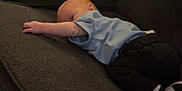 William is registered to the contest to win money with this photo: baby, infant, couch, sofa, clothing, blue_shirt, black_pants, socks, relaxing, indoor, furniture, pillow, home, child, person, lying_down, cute, small, resting, cozy