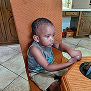 Elijah is registered to the contest to win money with this photo: child, boy, indoor, chair, table, wood, tile_floor, kitchen, cabinet, shorts, sleeveless_shirt, concentration, seated, furniture, young, portrait, casual, brown_skin, short_hair, home