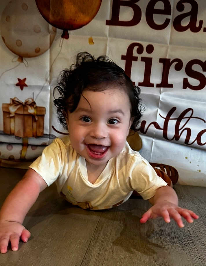 Naim joined the competition — help win amazing prizes! baby, child, smiling, crawling, curly_hair, birthday, balloons, presents, floor, wooden_floor, tshirt, happy, indoors, celebration, face, person, young_child, excited, background, decorations