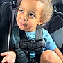 blue_shirt, car_interior, car_seat, casual_clothing, child, curly_hair, cute, indoors, looking_up, person, playful_expression, portrait, safety_harness, seat, seatbelt, sitting, toddler, vehicle, young_child, young_person