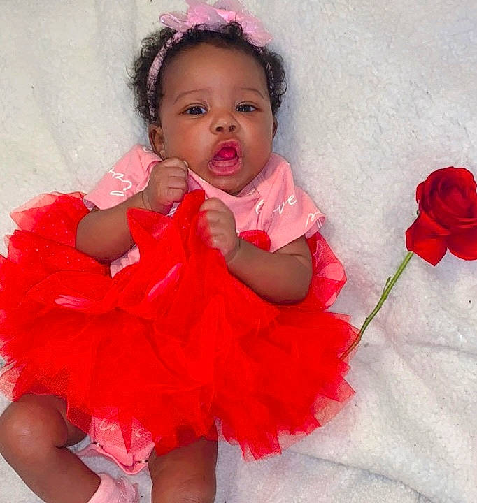 Emani is registered to the contest to win money with this photo: arm, baby_toddler_clothing, cheek, dress, eyelash, facial_expression, finger, flower, gesture, hand, happy, lip, organ, person, plant, product, skin, sleeve, standing, textile