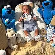 Zion is registered to the contest to win money with this photo: baby, infant, plush_toy, cookie_monster, teddy_bear, cushion, white_shoes, striped_overalls, milestone_marker, soft_lighting, smiling, indoor, cozy, cute, child, person, fabric, quilt, toy, comfortable