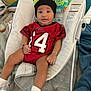 Zion is registered to the contest to win money with this photo: baby, infant, child, red_jersey, black_beanie, baby_rocker, blanket, socks, bracelet, indoor, soft_furniture, colorful_background, toy_pattern, relaxed, seated, cute, portrait, small_child, comfortable, cozy