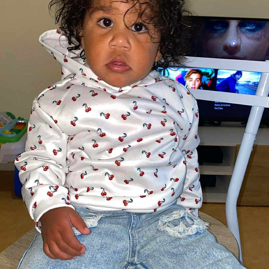 Assa participe au concours pour gagner de l'argent avec cette photo : casual_clothing, cherry_print, child, curious_expression, curly_hair, face, fashion, floor, hand, home_interior, indoor, person, ripped_jeans, screen, sitting, television, toddler, white_hoodie, wooden_chair, young_child