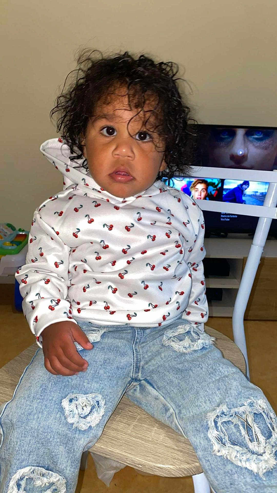 Assa participe au concours pour gagner de l'argent avec cette photo : toddler, child, curly_hair, white_hoodie, cherry_print, ripped_jeans, wooden_chair, indoor, television, screen, face, person, sitting, casual_clothing, young_child, curious_expression, home_interior, floor, hand, fashion