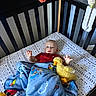 baby, crib, blanket, plush_toy, duck, toy, infant, child, sleeping_area, blue_blanket, red_clothing, playful, indoor, carpet, black_crib, patterned_sheet, hanging_toy, tongue_out, cute, young_child