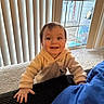toddler, child, smiling, indoors, hoodie, carpet, hand, vertical_blinds, glass_door, deck, window, young_child, person, happy, home, casual, clothing, face, teeth, curtains