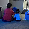 child, children, back_view, indoor, carpet, toy, living_room, playtime, home, furniture, refrigerator, cabinet, person, baby, sitting, floor, casual_clothing, glasses, household_items, family