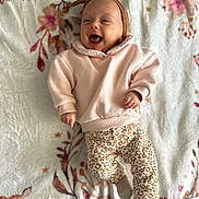 Azélia a rejoint le concours — aidez-le/la à gagner de superbes lots ! adorable, animal_print_pants, baby, blanket, child, cozy, cute, floral_pattern, happy, headband, indoors, infant, lying_down, pink_sweater, playful, portrait, smiling, socks, soft_texture, warm_colors
