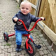 Logan is registered to the contest to win money with this photo: toddler, child, tricycle, outdoor, pavement, smiling, hoodie, jeans, footwear, fence, toy, vehicle, person, happy, young_child, play, casual_clothing, daylight, blue_eyes, cute