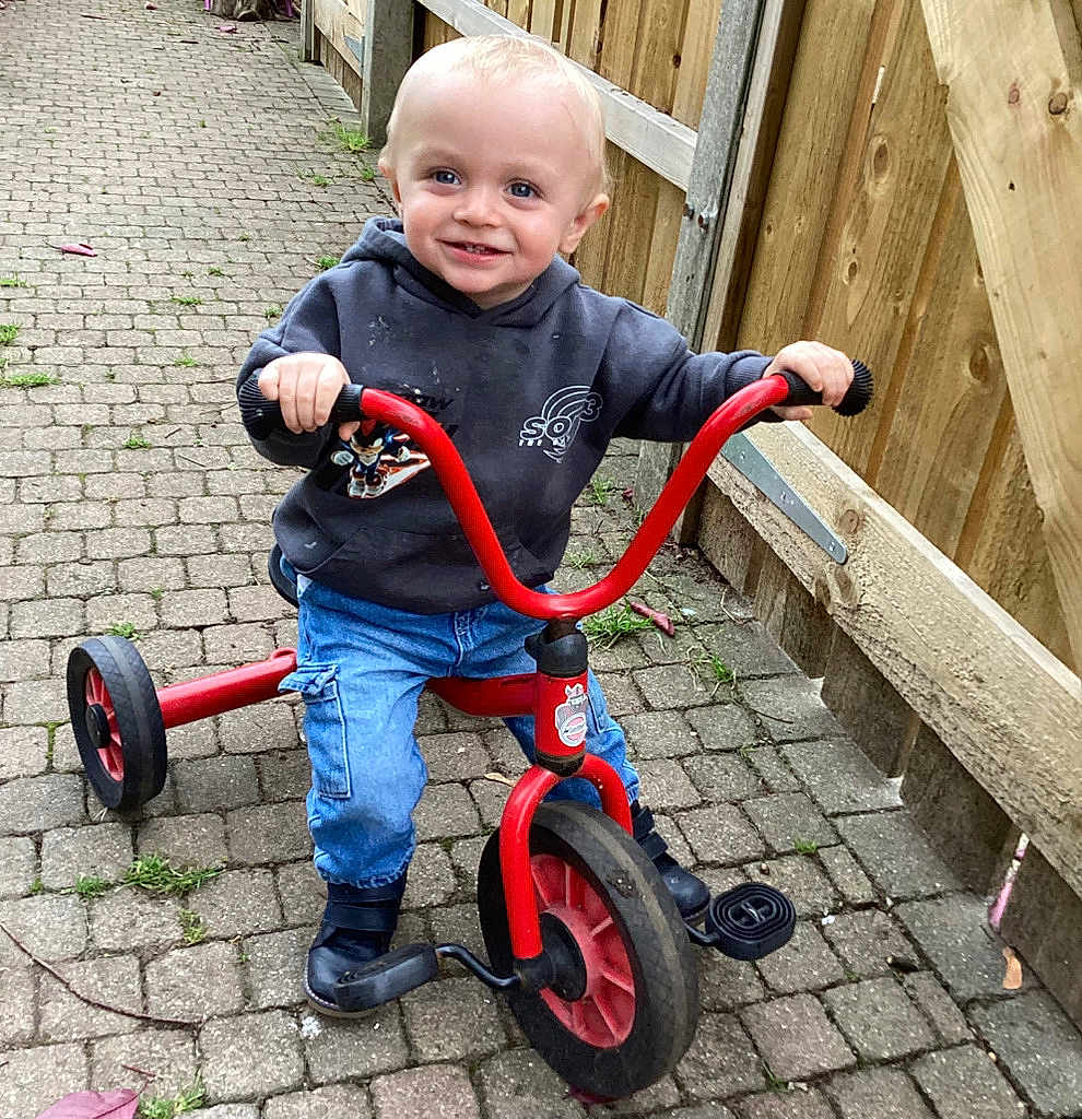 Logan is registered to the contest to win money with this photo: toddler, child, tricycle, outdoor, pavement, smiling, hoodie, jeans, footwear, fence, toy, vehicle, person, happy, young_child, play, casual_clothing, daylight, blue_eyes, cute