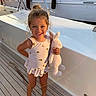barefoot, boat, bunny, child, cute, girl, hair_bun, happy, nautical, outdoor, playful, smiling, stuffed_animal, summer, sunlight, sunset, swimsuit, toy, vacation, wooden_deck