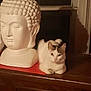 cat, white_cat, buddha_sculpture, indoor, wooden_surface, statue, calm, pet, animal, decor, brown_spots, feline, quiet, resting, household, table, figurine, still_life, soft_light, window