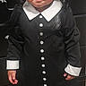 child, toddler, costume, black_dress, white_collar, braided_hair, barefoot, carpet, indoor, serious_expression, portrait, person, standing, dresser, furniture, buttons, long_sleeves, hair, cute, young_child
