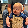 baby, child, infant, overalls, whale_pattern, blue_shirt, adult_supporting, indoor, wooden_floor, blurred_background, cute, focused_gaze, clenched_fists, portrait, person, casual_clothing, light_skin, short_hair, happy, seated_adult