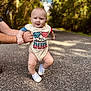 baby, child, infant, person, outdoor, road, trees, greenery, sunlight, shoes, onesie, happy, smiling, adult_hand, holding, walking, nature, blurred_background, cute, patriotic