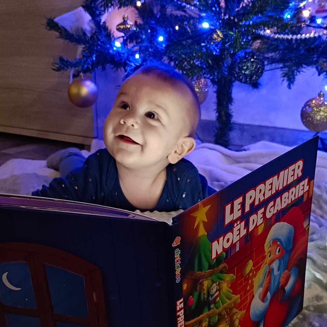 Gabriel a rejoint le concours — aidez-le/la à gagner de superbes lots ! baby, blanket, book, celebration, child, christmas_tree, cute, decorations, face, festive, happy, holiday, indoors, lights, ornaments, present, reading, smile, socks, winter