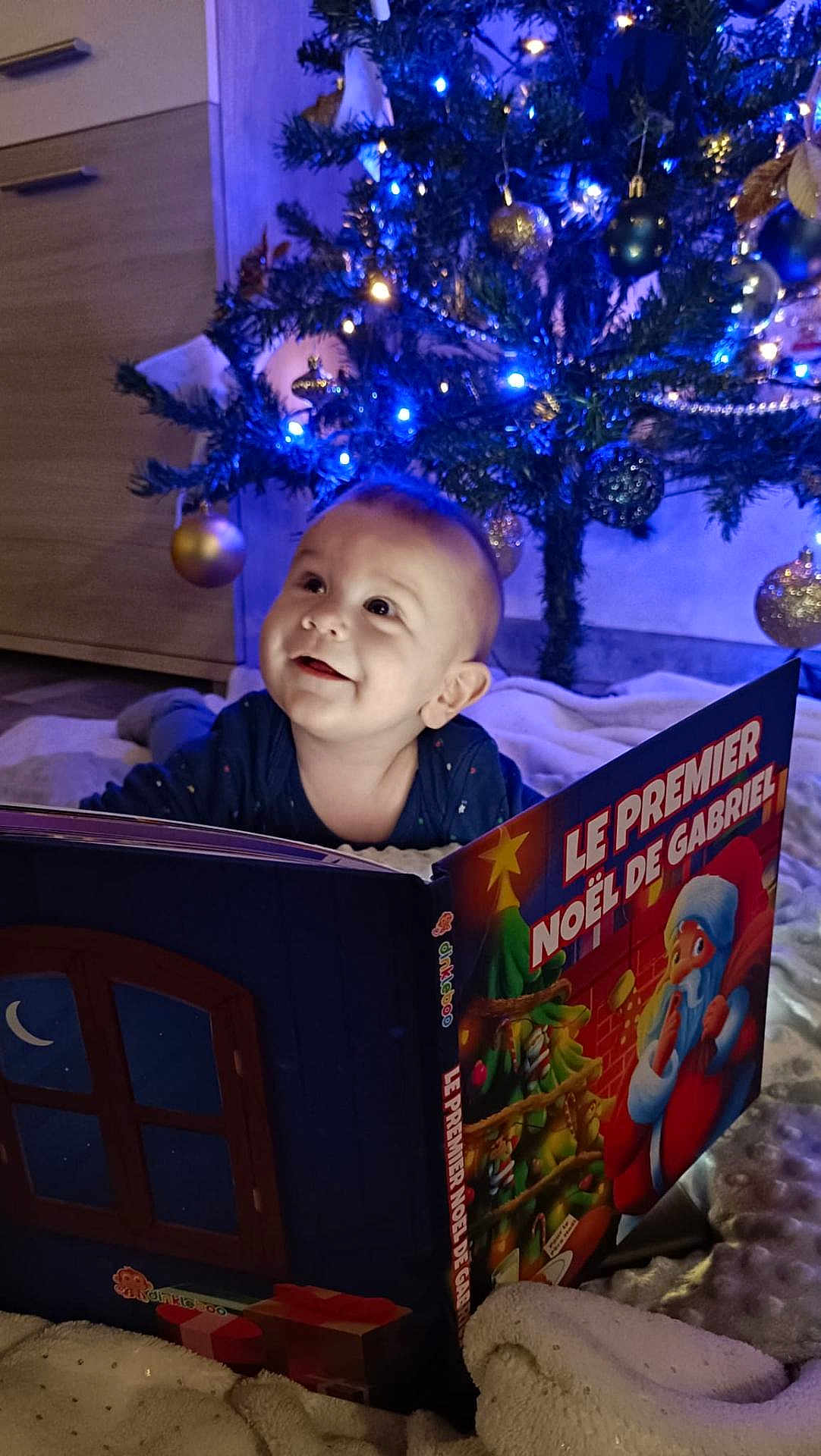 Gabriel a rejoint le concours — aidez-le/la à gagner de superbes lots ! baby, child, christmas_tree, book, holiday, lights, ornaments, blanket, smile, indoors, festive, decorations, cute, happy, reading, winter, socks, face, present, celebration