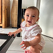 Solea participe au concours pour gagner de l'argent avec cette photo : baby, child, smiling, white_outfit, red_trim, squatting, floor, tiled_floor, hand, foot, table_leg, furniture, wooden_furniture, indoor, happy, person, cute, infant, face, bright