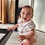 baby, child, smiling, white_outfit, red_trim, squatting, floor, tiled_floor, hand, foot, table_leg, furniture, wooden_furniture, indoor, happy, person, cute, infant, face, bright