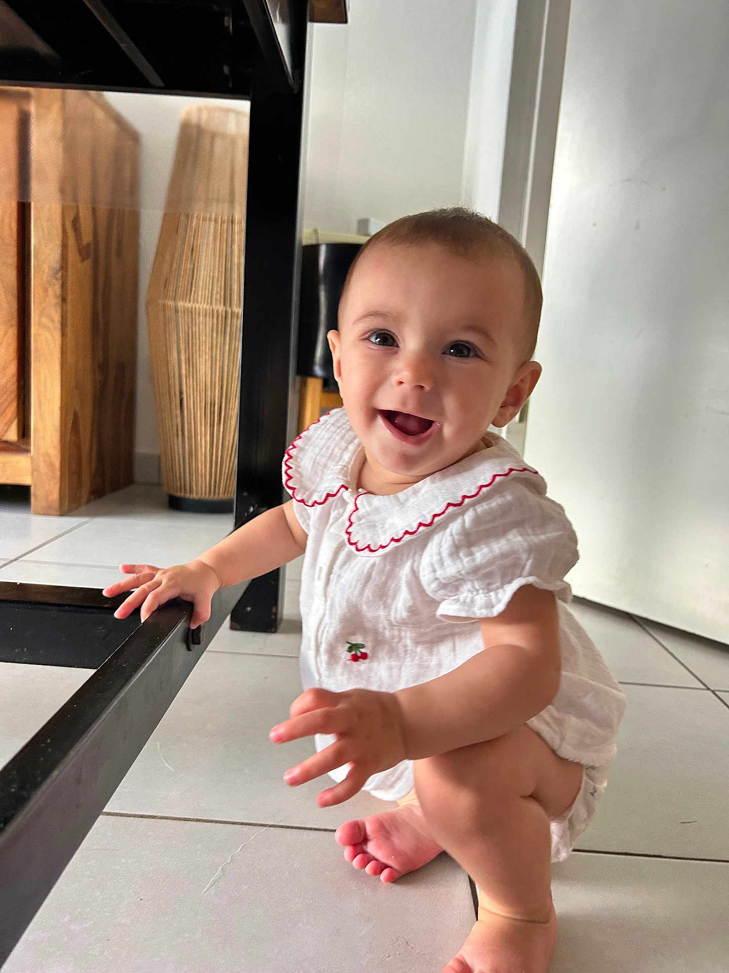 Solea participe au concours pour gagner de l'argent avec cette photo : baby, child, smiling, white_outfit, red_trim, squatting, floor, tiled_floor, hand, foot, table_leg, furniture, wooden_furniture, indoor, happy, person, cute, infant, face, bright