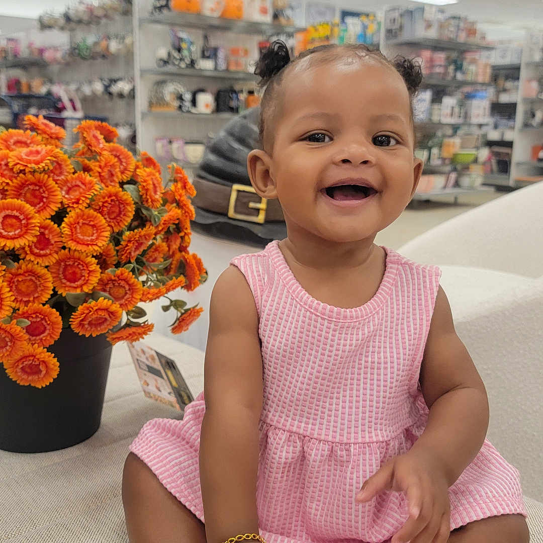 Jurnee is registered to the contest to win money with this photo: baby, bracelet, child, couch, cute, flowers, happy, home_goods, indoor, infant, orange_flowers, person, pigtails, pink_dress, portrait, pot, seated, smiling, socks, store