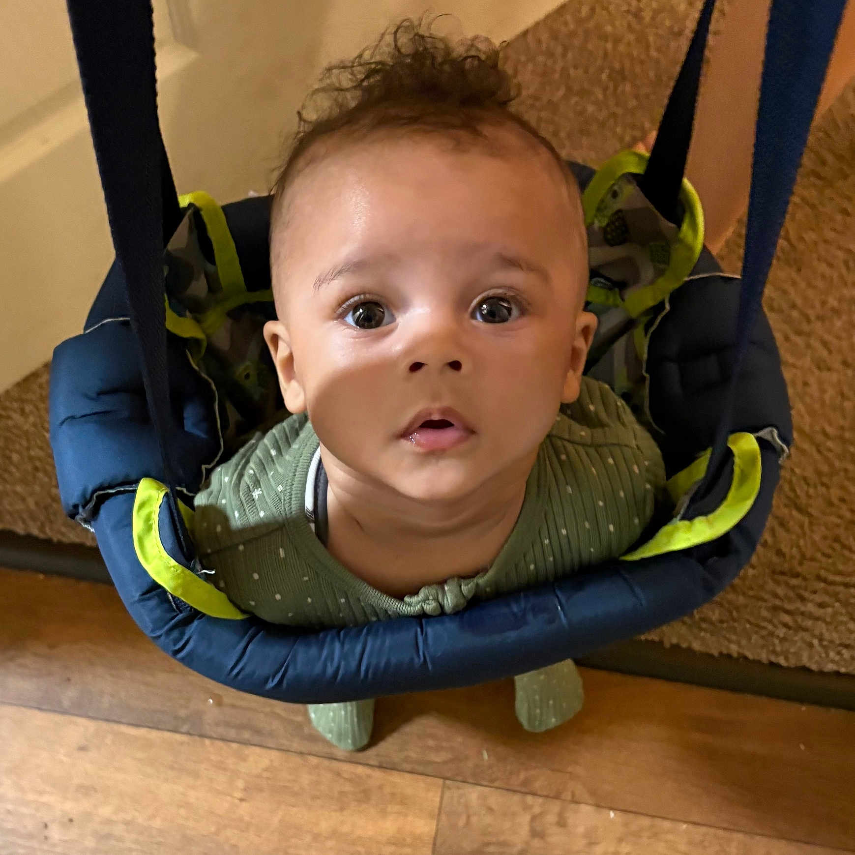 Damari is registered to the contest to win money with this photo: baby, child, infant, face, eyes, curly_hair, baby_jumper, indoor, carpet, wood_floor, clothing, green_outfit, looking_up, cute, young_child, expression, person, closeup, sitting, play
